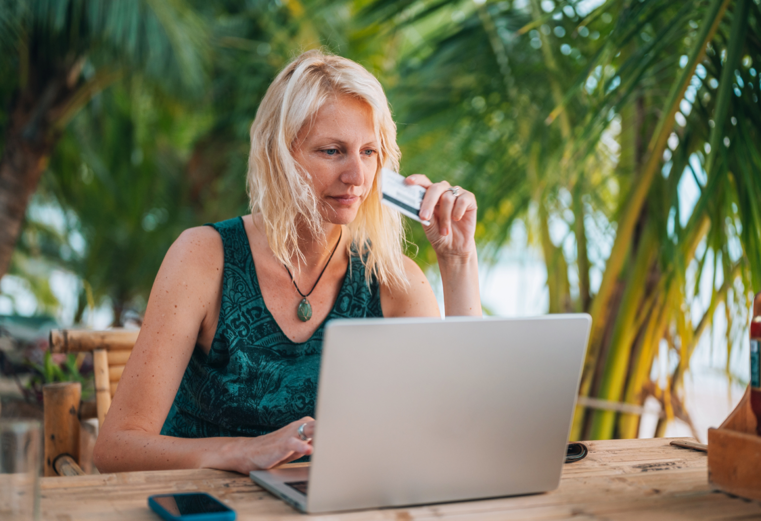 blonde woman holding credit card while shopping on laptop in a tropical setting