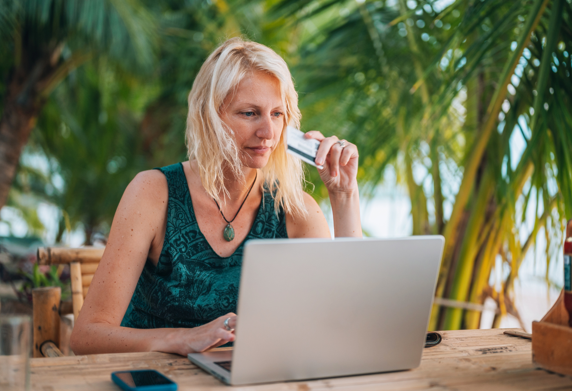 blonde woman holding credit card while shopping on laptop in a tropical setting